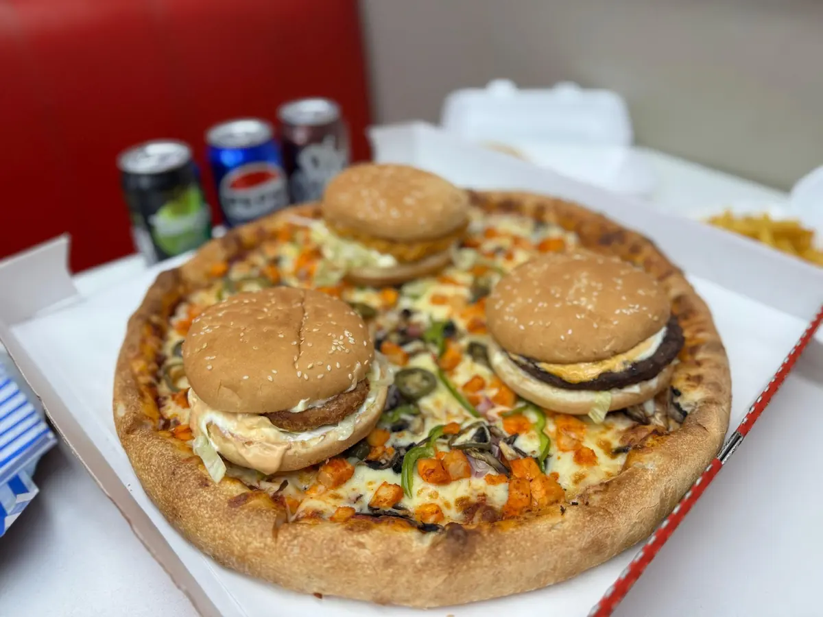 A loaded pizza topped with two burger buns in an open box, drinks behind